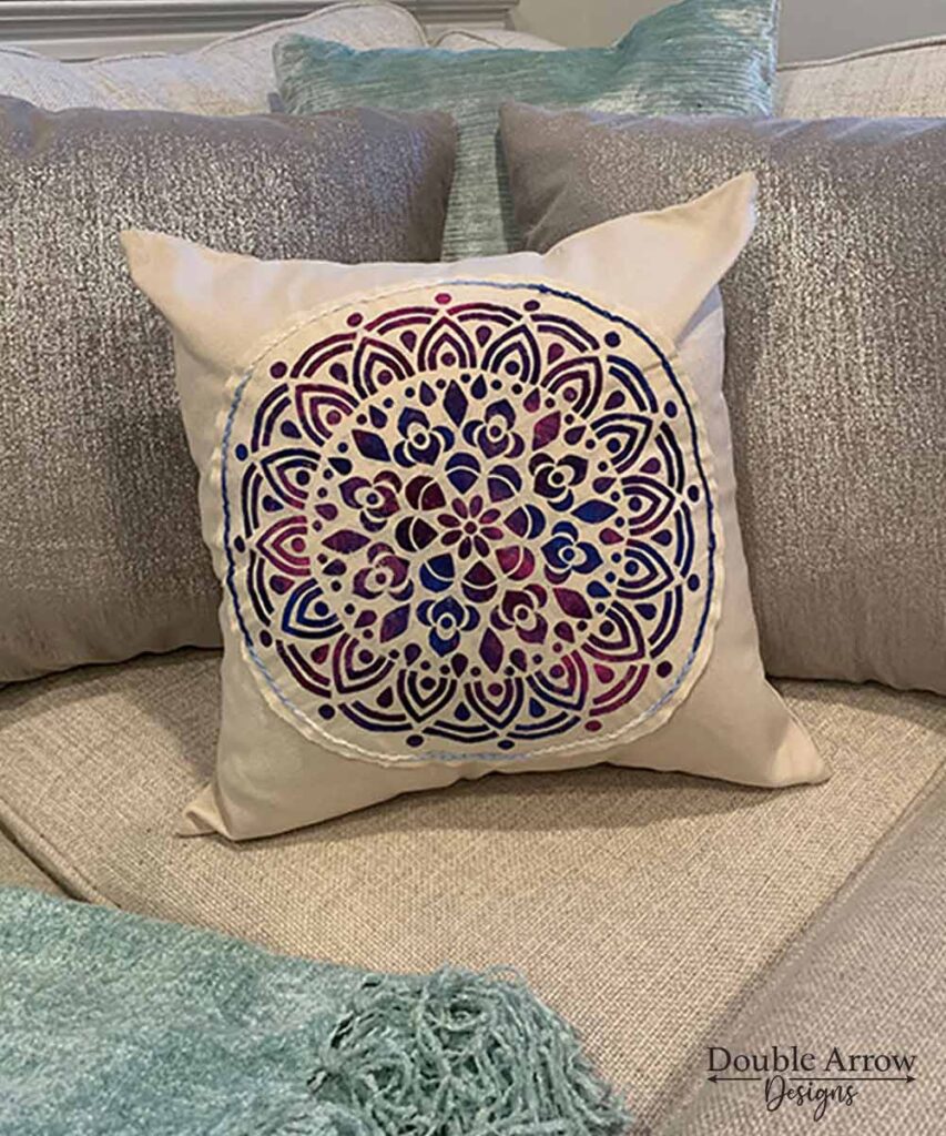 medallion- stencilled pillow on a cream colored couch with other pillows behind it.