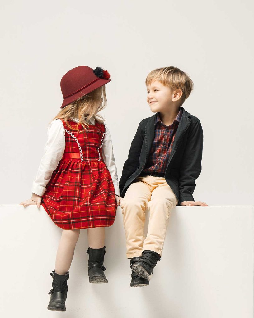 A girl dressed in a red plaid dress looking at a smiling boy in a blazer and tan pants both sitting