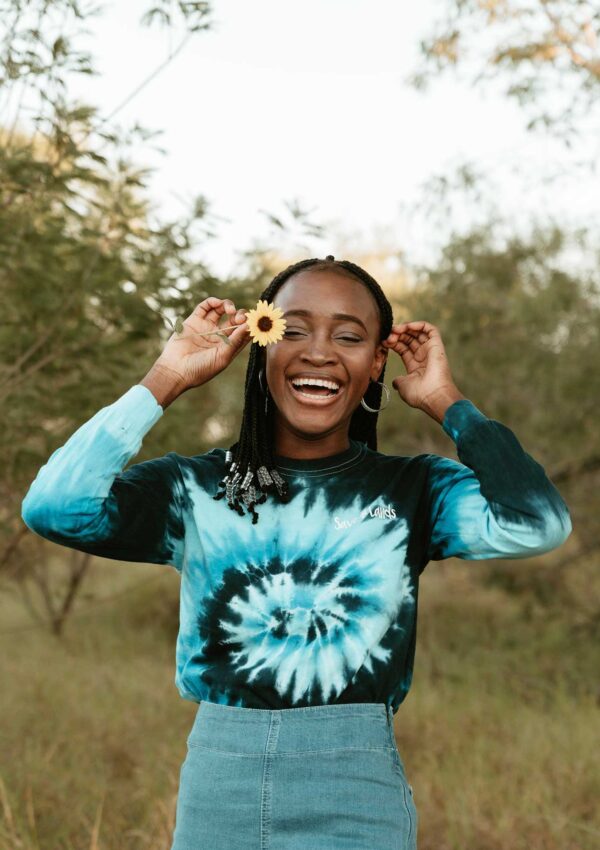 woman wearing a blue swirl tie dye tee outside laughing holding up a yellow flower