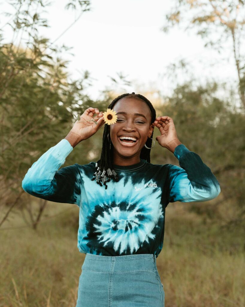 woman wearing a blue swirl tie dye tee outside laughing holding up a yellow flower