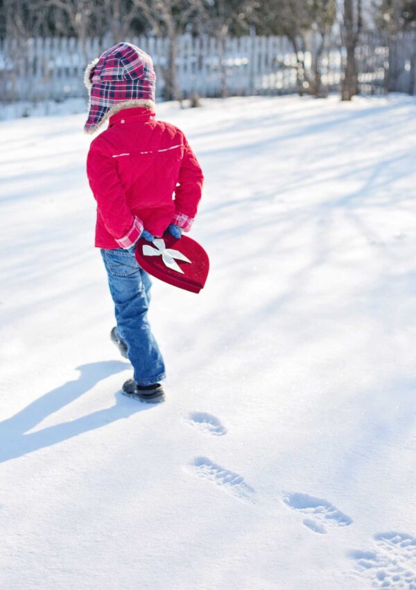 toddler boy in plaid hat walking away in the snow holding a heart box of valentines day candies