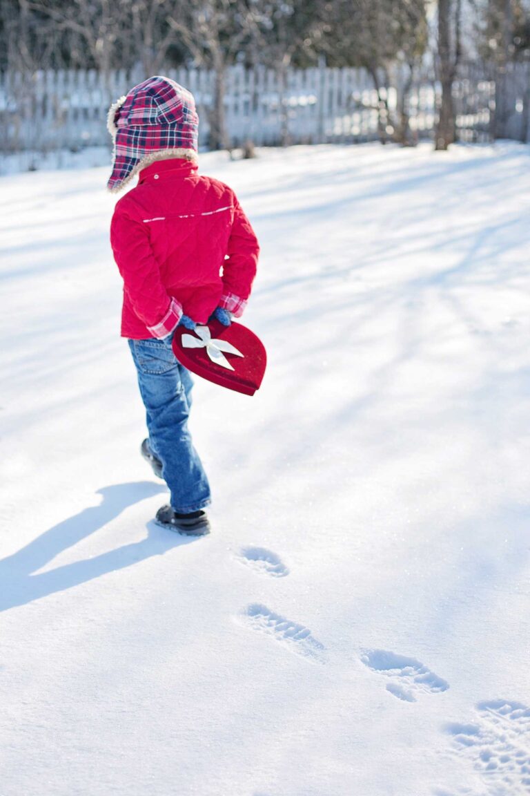 toddler boy in plaid hat walking away in the snow holding a heart box of valentines day candies