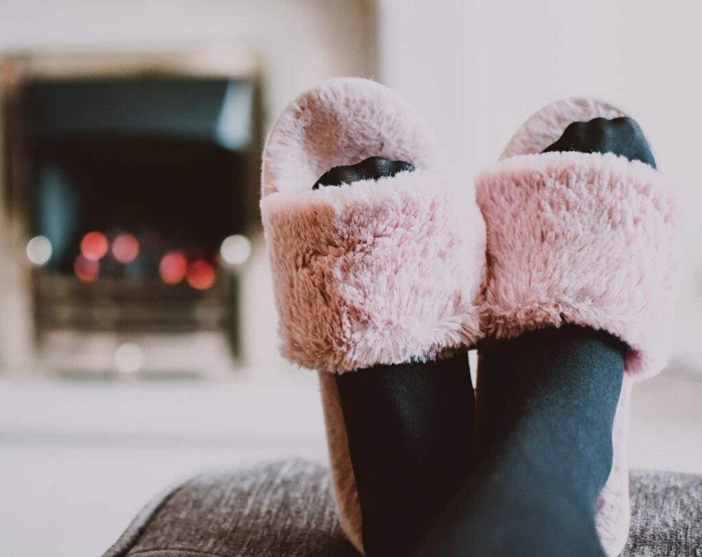 Best Slippers To Wear At Home. pink fuzzy slippers on black socked feet  in front of a fireplace