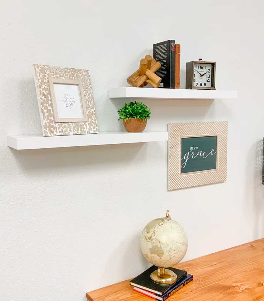 two white floating shelves with frame, books, clock plant and wooden object.