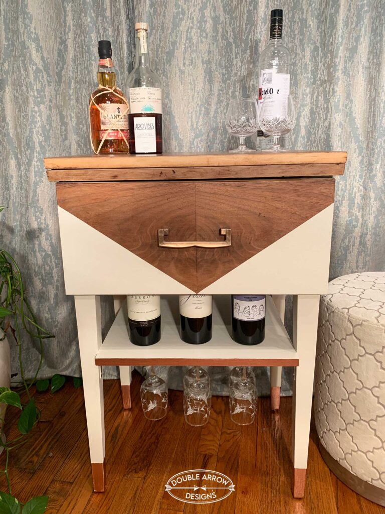 Repurposed sewing cabinet into a bar cart. Wood with cream painted legs . Bottles of liquor and glasses on display.