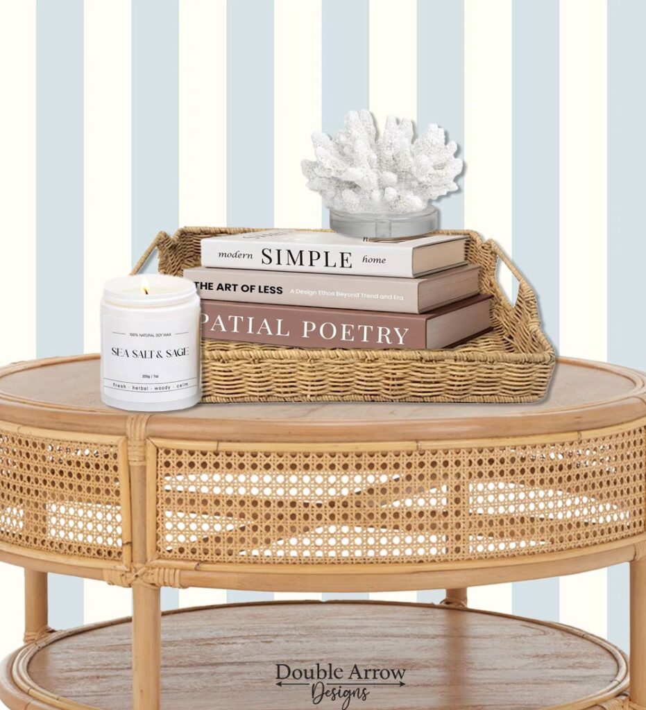coastal decor on round coffee table, rattan tray, book stack, faux resin coral, sea and salt sage candle in front of a blue and white striped wall
