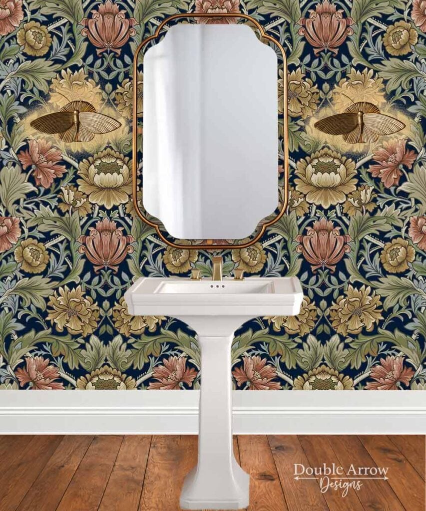 Moody powder room with vintage floral wallpaper, brass framed mirror, white pedestal sink, warm wood flooring, and brass accents.
